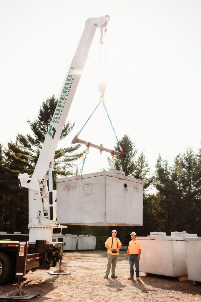 Two workers standing near a large precast concrete septic tank being lifted by a boom truck from Superior Precast Concrete, located in Cable, Wisconsin. The scene is set outdoors at their concrete production facility surrounded by pine trees.